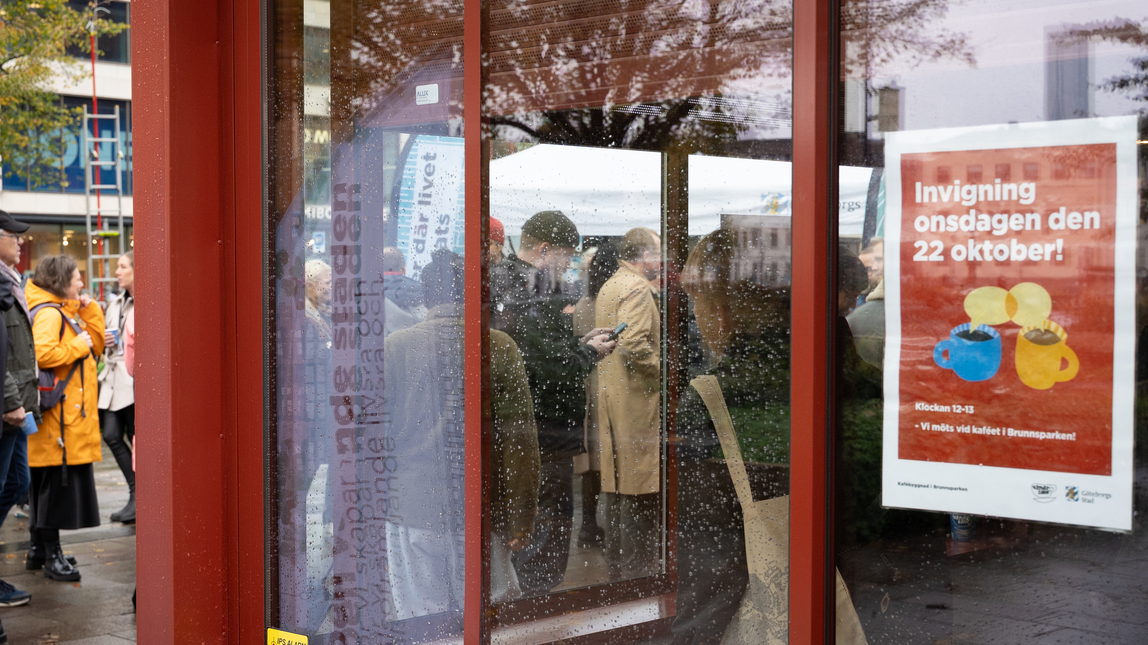 Personer syns genom regniga glasrutor. P&aring; ena glasrutan syns en affisch med texten Invigning onsdagen den 22 oktober.
