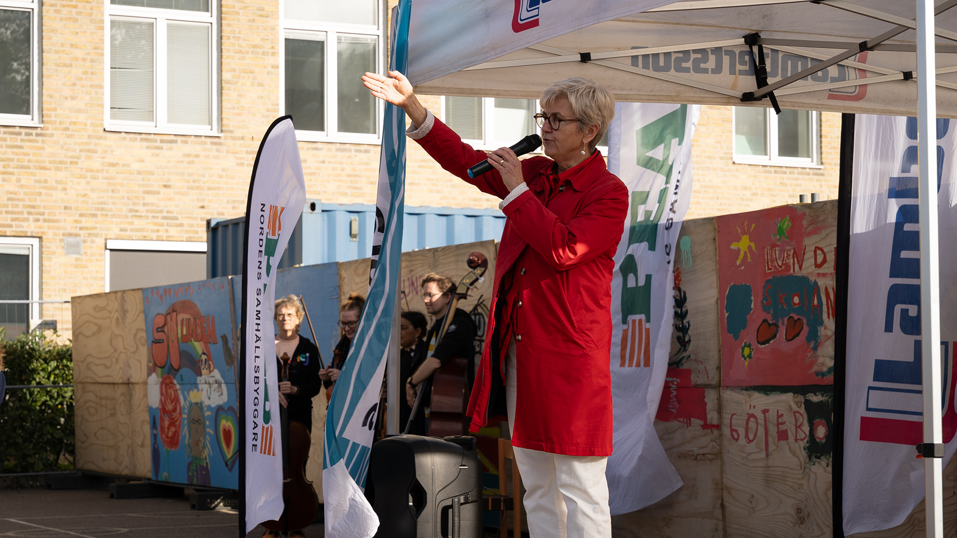 En kvinnar i röd kappa med mikrofon i handen som gestikulerar mot publiken.