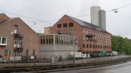 En rödbrun tegelbyggnad i fyra våningar, med en mindre grå träbyggnad framför.