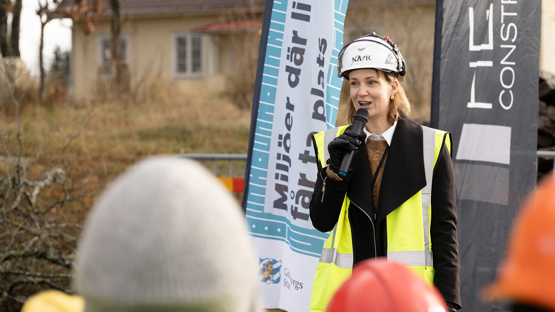 En kvinna i varselv&auml;st och bygghj&auml;lm st&aring;r och pratar i en mikrofon.