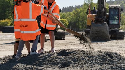 Tre barn i för stora reflexvästar gräver med en treskaftad spade. I bakgrunden en grävmaskin.