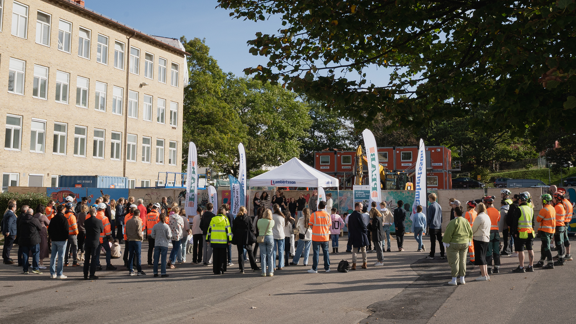 Många människor på en skolgård framför ett byggplank och en tillfällig scen med tälttak.