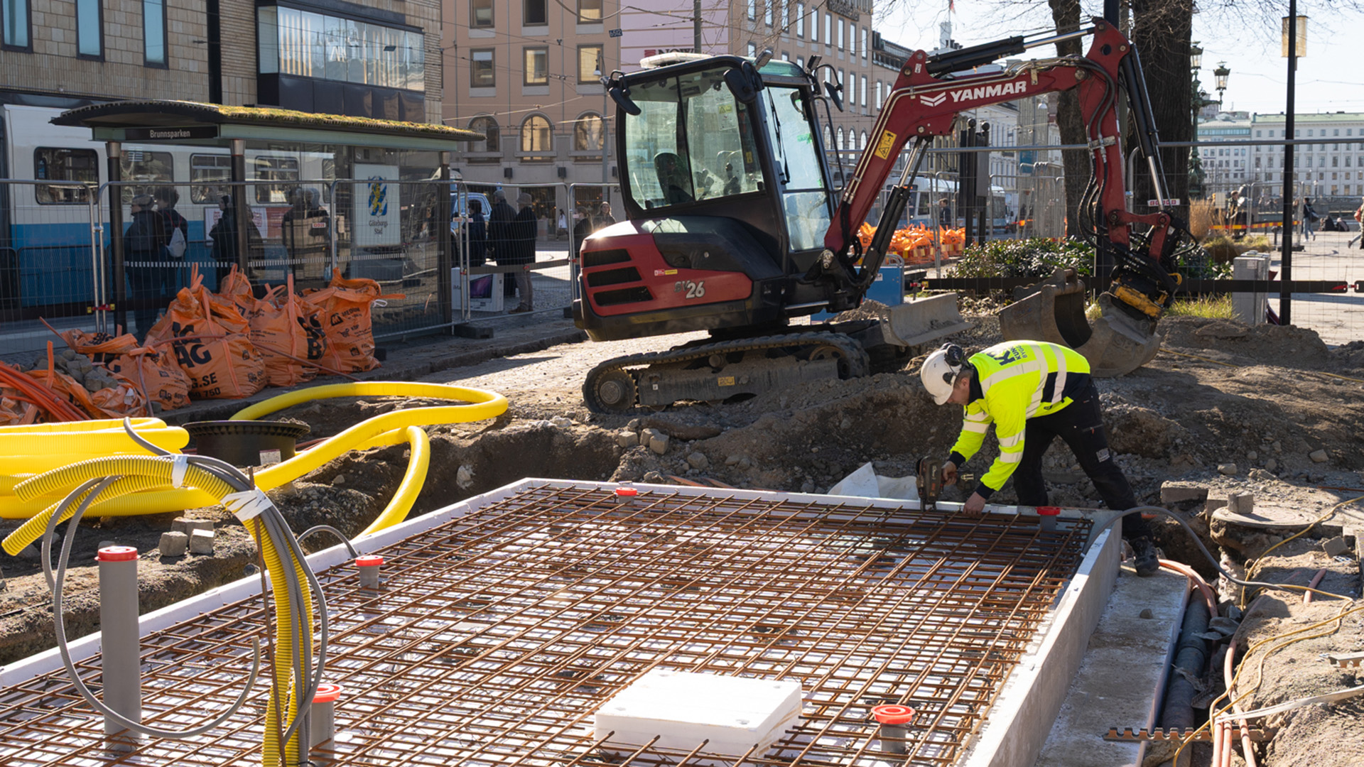 En person i varselkläder grejar med armeringsnät i en gjutform. I bakgrunden står en grävamaskin. En spårvagn kör förbi.
