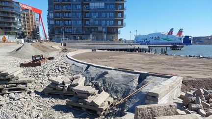 Sten, grus och jord vid älven där en park byggs. I bakgrunden två bostadshus i sex våningar och Eriksbergskranen.