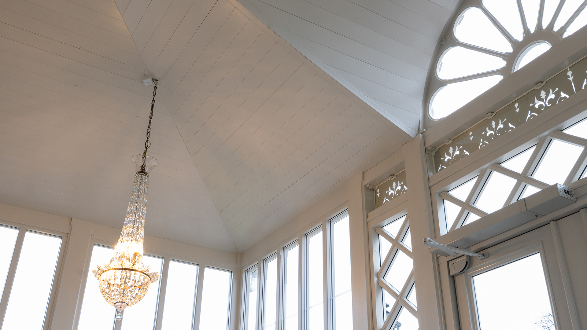Ett vitt innertak i tr&auml;panel med m&aring;nga vinklar och vr&aring;r, dekorerade f&ouml;nster och en kristallkrona.