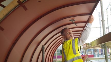 En person i varselkläder står på en byggnadsställning under ett böjt tak.