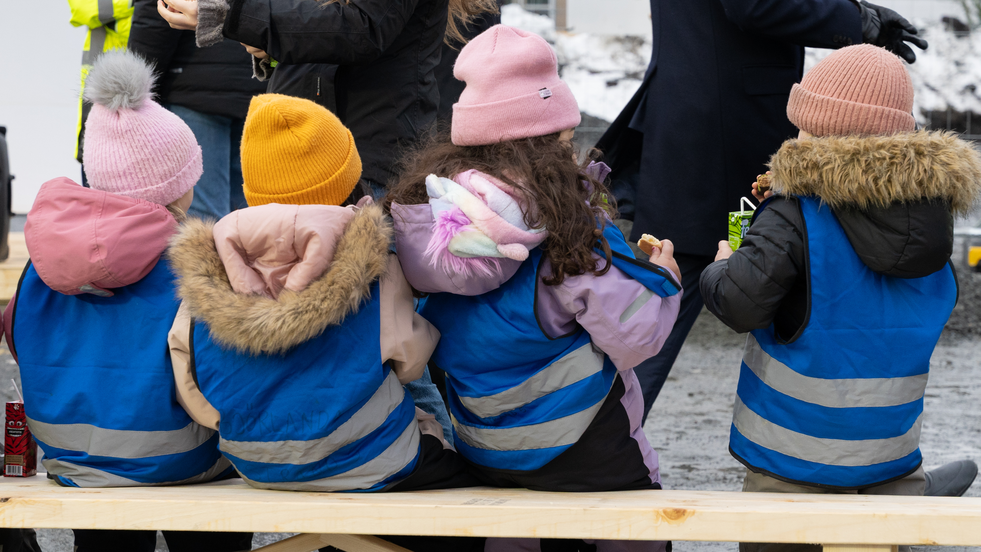 Fyra barn i bl&aring; reflexv&auml;star sitter p&aring; en tr&auml;b&auml;nk med ryggen mot kameran.