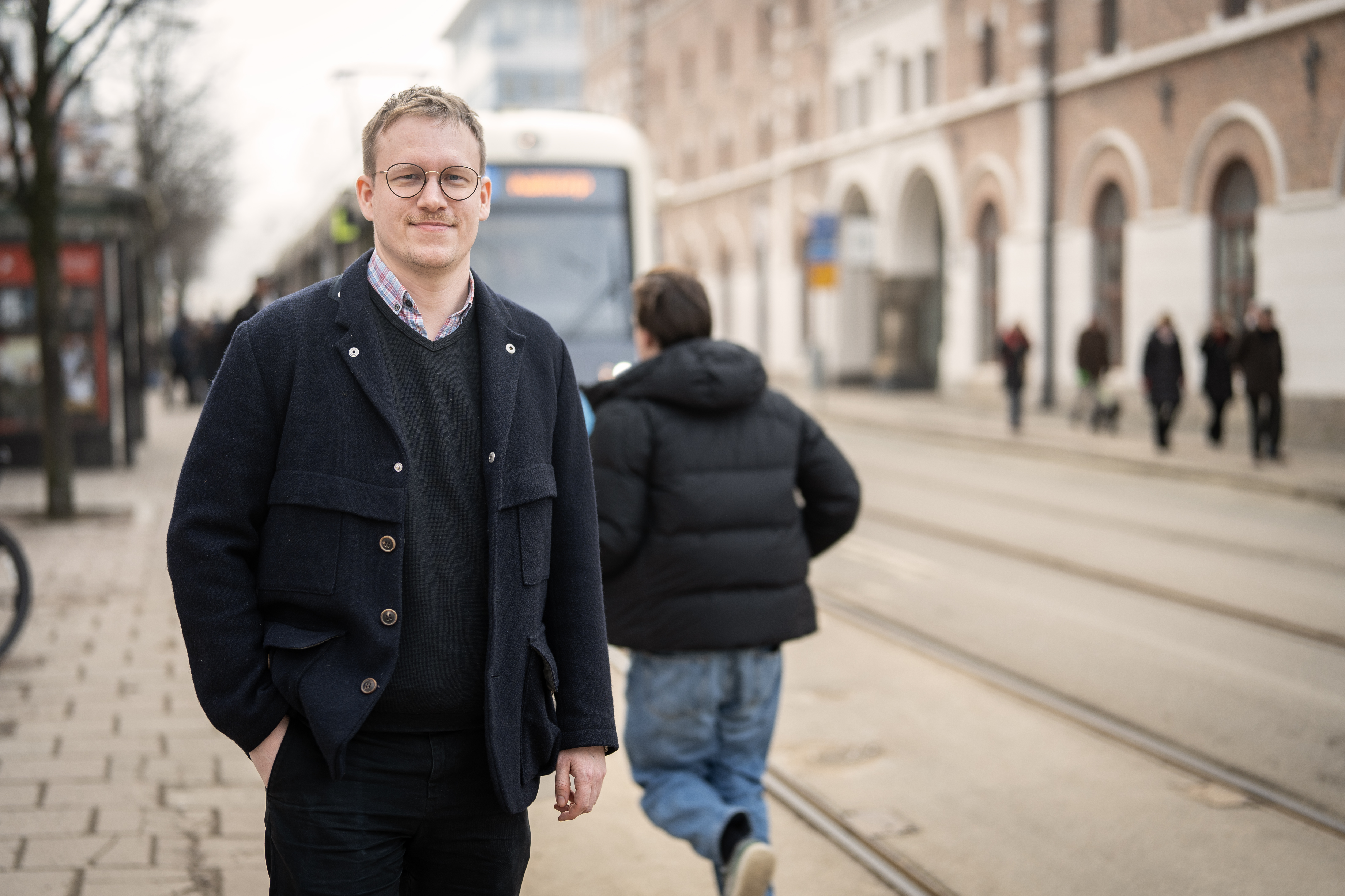 En man står framför en spårvagn, vänd mot kameran. I bakgrunden syns en annan man springa för att hinna med vagnen.
