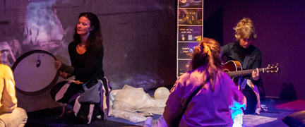 Personer sitter på golvet i ett rum med sinnestimulerande material. En person spelar gitarr och en annan spelar trumma. Man ser ryggen på två personer som deltar i musikdramat.