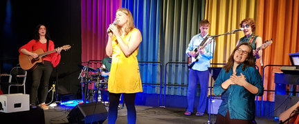 Flera personer står på en scen. De spelar musikinstrument och sjunger.