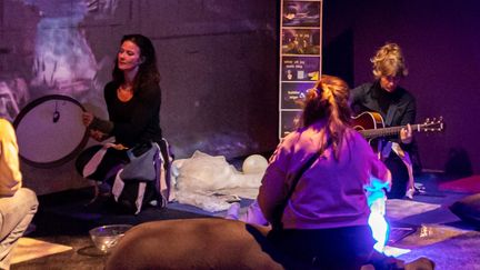 Personer sitter på golvet i ett rum med sinnestimulerande material. En person spelar gitarr och en annan spelar trumma. Man ser ryggen på två personer som deltar i musikdramat.