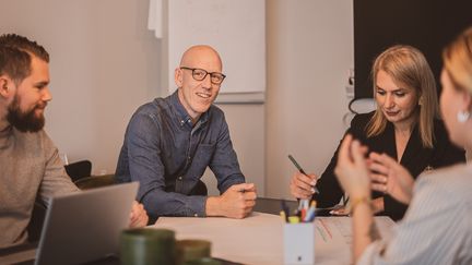 Ett pågående möte i ett mötesrum mellan 4 personer. I bakgrunden syns en TV-skärm och en tavla med uppsatta post it-lappar. 