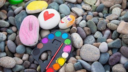 Massor av släta stenar varav några är målade. En palett med vattenfärger ligger på stenarna.