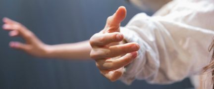Närbild på en hand i fokus som är i rörelse. Iklädd en vit fladdrig skjorta. Två personer dansar på scen och man ser den andra personens hand suddigt i bakgrunden.