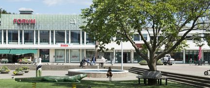 Vy över Kortedala Forum-byggnaden intill Kortedala Torg. På torget finns det grönskande träd och en fontän, där människor strosar runt.