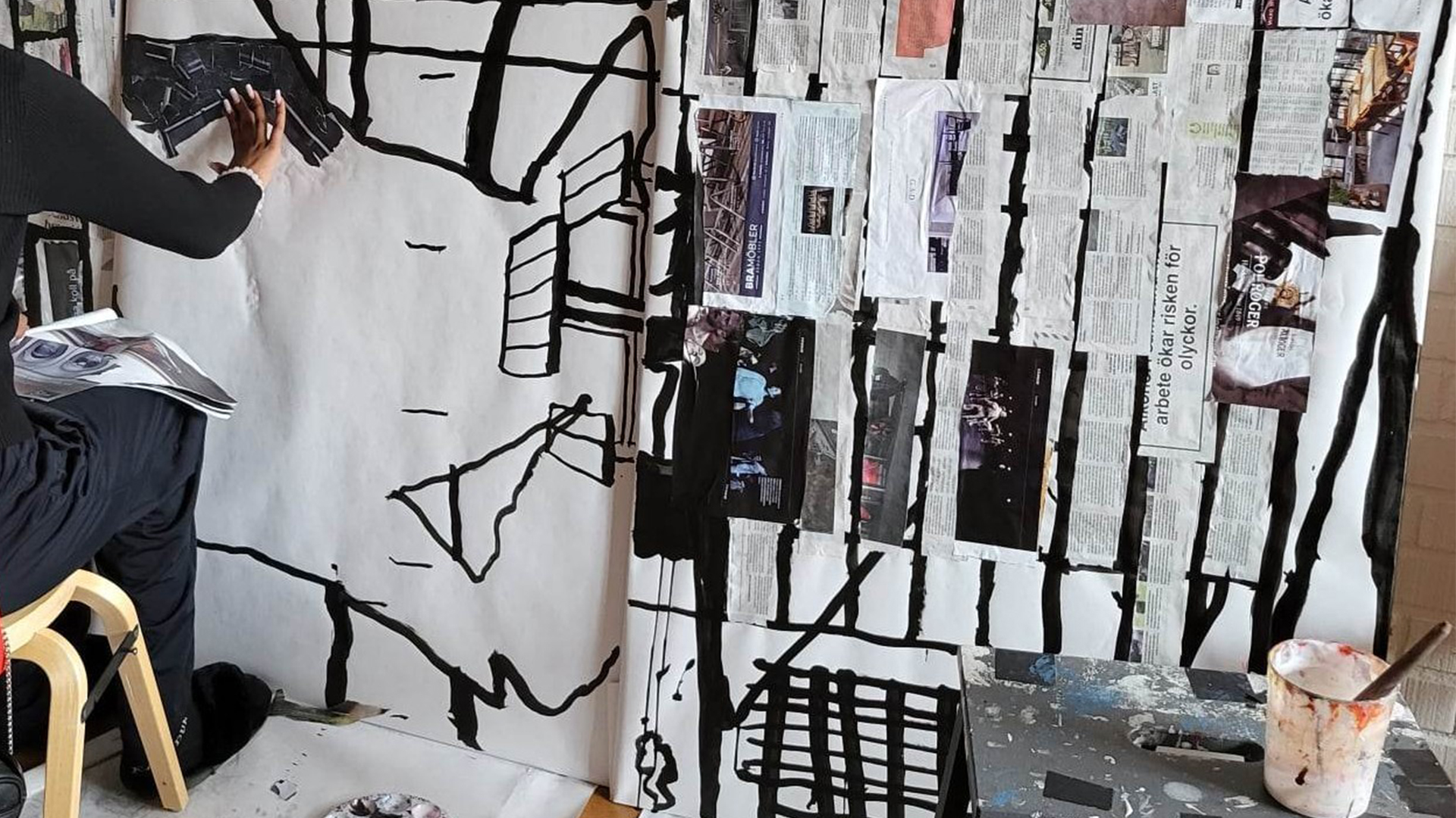 Konstnär i svarta kläder, sittande på stol, syns i vänsterkanten men vi ser bara armen, handen och benet. Hen målar svarta former på vit duk.
