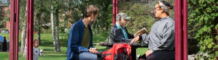 Tre personer runt ett bord med bänkar utomhus. I bakgrunden syns ett barn.