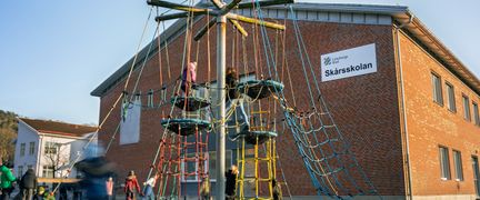 Några barn som klättrar och leker i en lekställning på en skolgård framför en skolbyggnad med texten Skårsskolan.