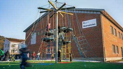 Några barn som klättrar och leker i en lekställning på en skolgård framför en skolbyggnad med texten Skårsskolan.