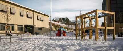 En snötäckt lekplats utanför Bergsgårdsskolan med en klätterställning och några skolbyggnader i bakgrunden.