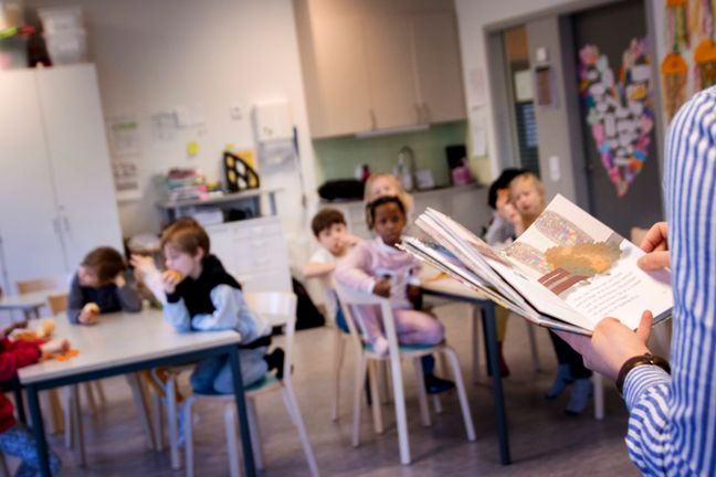 Yngre elever sitter på sina platser i klassrummet och lyssnar när deras lärare läser.