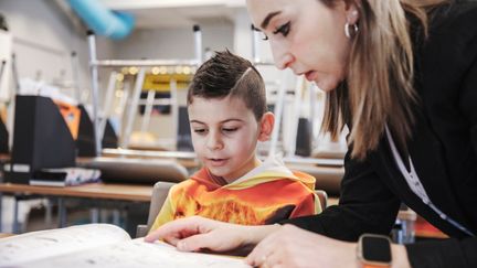 Lärare sitter vid skolbänken tillsammans med en elev och tittar i en bok.