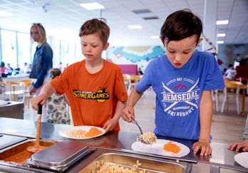 Elever tar mat i skolmatsalen En lärare står i bakgrunden.