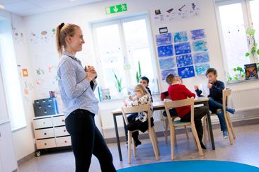 En glad lärare berättar för eleverna om något. Eleverna sitter runt bord i klassrummet. 