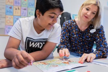 Undervisning i klassrummet. En lärare sitter bredvid en elev i skolbänken och pekar i en bok.