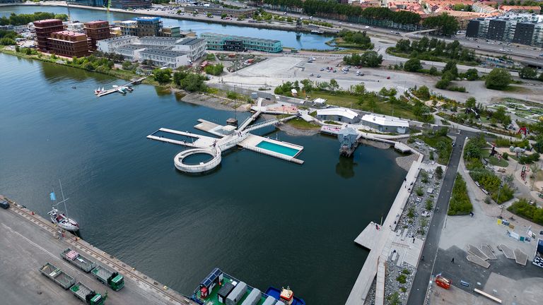 Tre flytande bassänger i ett hamnområde. På land omkring badet finns en park och en lekplats.