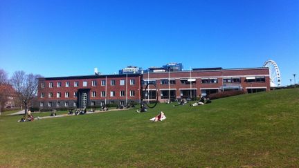 Bild på en av Göteborgs Universitets byggnader i närheten av Liseberg. Framför finns en grön gräsmatta där några sitter och njuter av solen.
