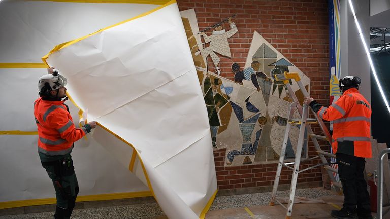 Två personer i varseljackor drar av stora pappers från en tegelvägg, så att Lisa Larssons konstverk kikar fram.