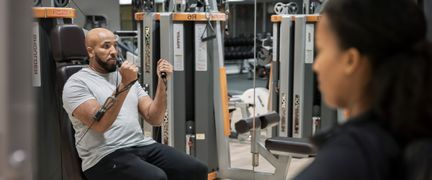 Person använder styrkemaskin. En till person skymtar i förgrunden