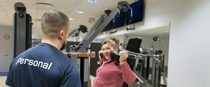 En person tränar i en gymmaskin och pratar med en person med texten Personal på ryggen.