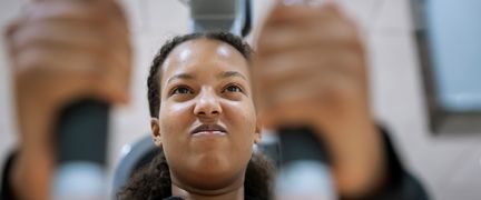 Ansiktet på en ung person som tar i ordentligt i en styrkemaskin. I förgrunden syns personens händer som håller i en maskinen