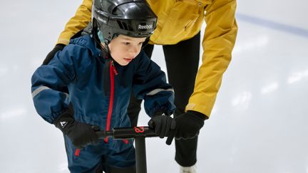 Ett barn åker skridskor med åkstöd. En vuxen i bakgrunden hjälper till.