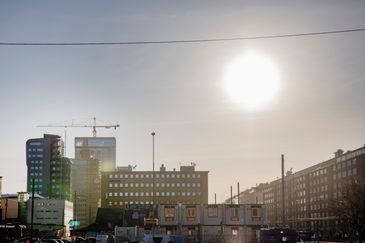 Solen skiner starkt i stadsmiljö. Mellan byggnaderna syns lyftkranar.
