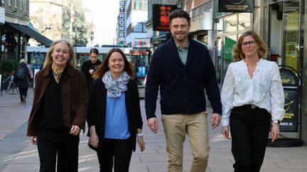 Fyra tjänstepersoner går leende mot kameran i stadsmiljö. I bakgrunden åker en spårvagn förbi.