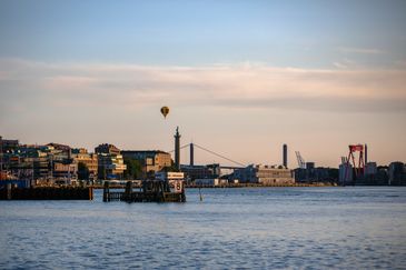 Utsikt från hamnkanten över Göteborgs Stads silhuett. En luftballong svävar över staden samtidigt som solen har börjat gå ner och nyansera den blåa himlen med rosa inslag.
