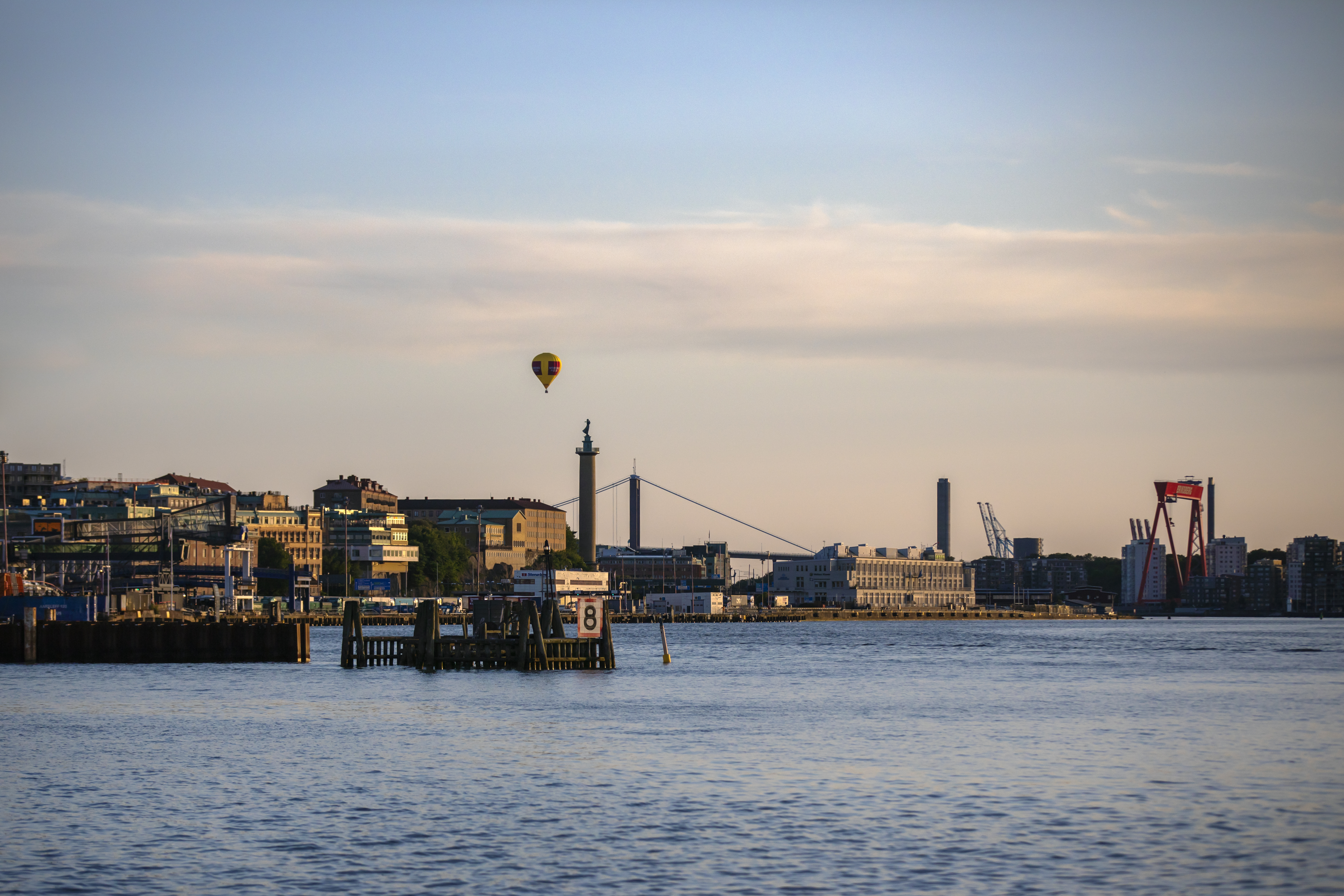 Utsikt från hamnkanten över Göteborgs Stads silhuett. En luftballong svävar över staden samtidigt som solen har börjat gå ner och nyansera den blåa himlen med rosa inslag.