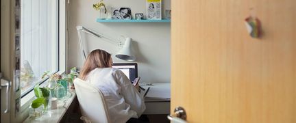 En ung person sitter och kollar mobilen framför sin laptop i ett ombonat rum.