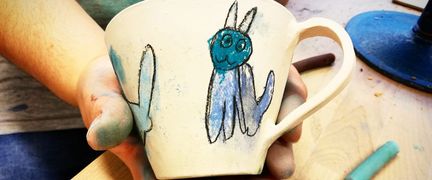 En hand håller i en handmålad mugg som föreställer en blå katt.