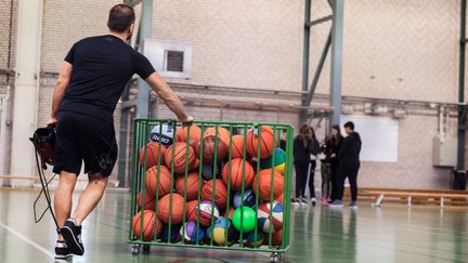 En man med en korg med många bollar samt barn i en idrottshall.