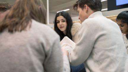 En grupp ungdomar sitter vid ett bord och samarbetar, där en ung kvinna tittar in i kameran och ler. 