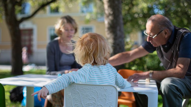 Bild på två vuxna och ett barn som sitter runt ett bord och spelar spel i en trädsgård.
