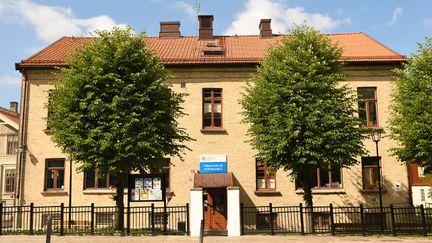 En gul tegelbyggnad med röda fönster. Två träd med gröna löv står framför huset.