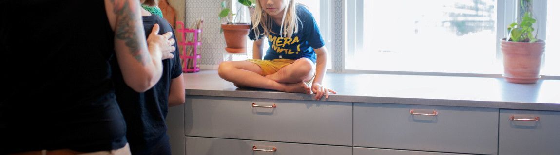 Ert barn med gula shorts sitter med benen i kors på en grå köksbänk. Till vänster i bild ser man ryggen av en vuxen som har ett svart linne på sig, hen håller armen om ett barn som lagar mat.