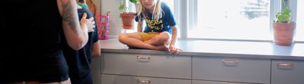 Ert barn med gula shorts sitter med benen i kors på en grå köksbänk. Till vänster i bild ser man ryggen av en vuxen som har ett svart linne på sig, hen håller armen om ett barn som lagar mat.
