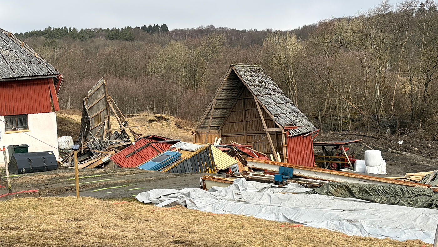En röd, delvis sammanrasad ladugårdsbyggnad med utspridda tak- och väggdelar på mark som har rört sig efter ett jordskred