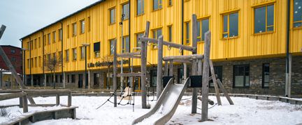 En skolgård med en rutschkana och klätterställning framför en gul skolbyggnad.